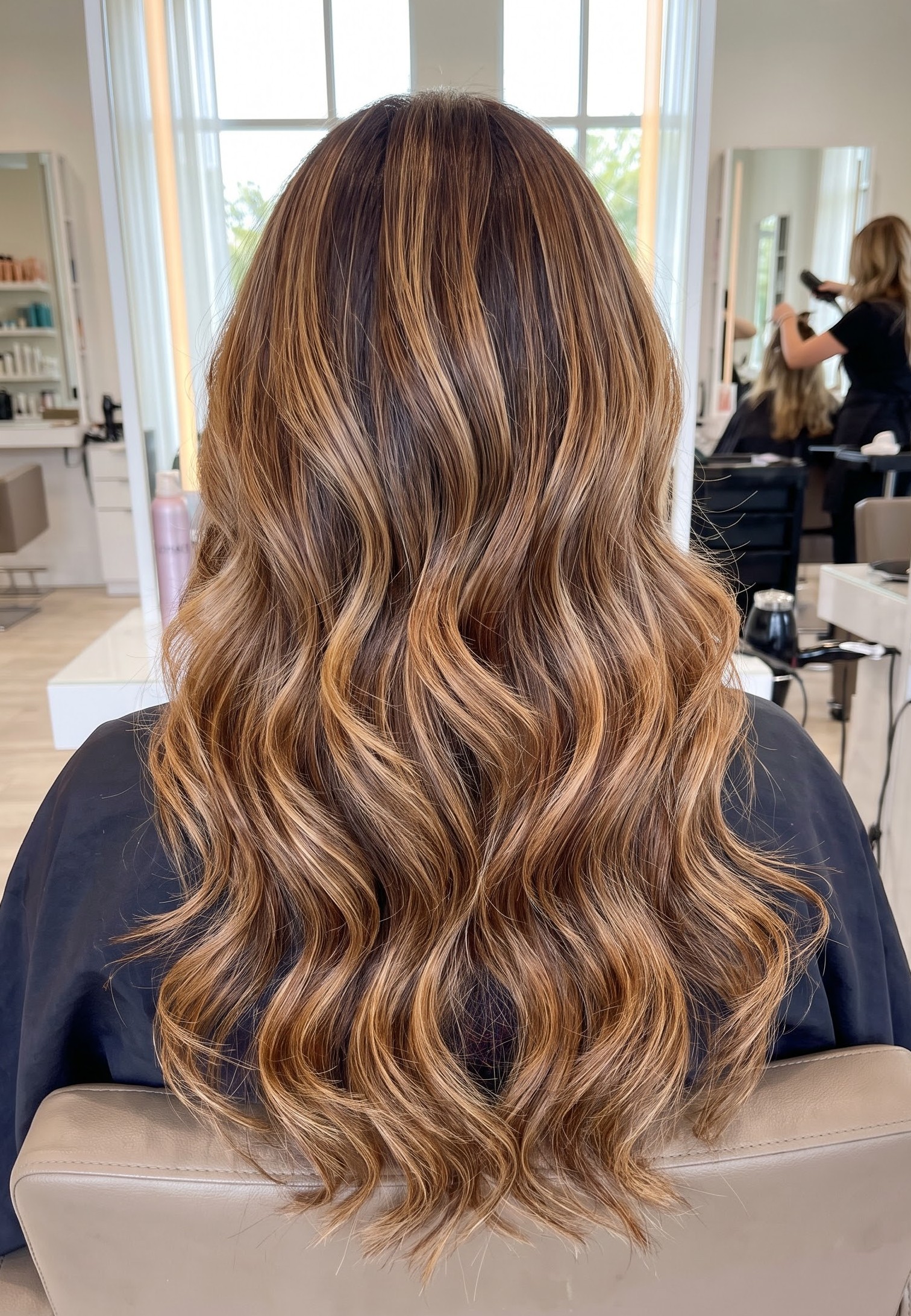 Brunette hair with caramel balayage highlights styled in loose curled layers beside a bright arched salon window