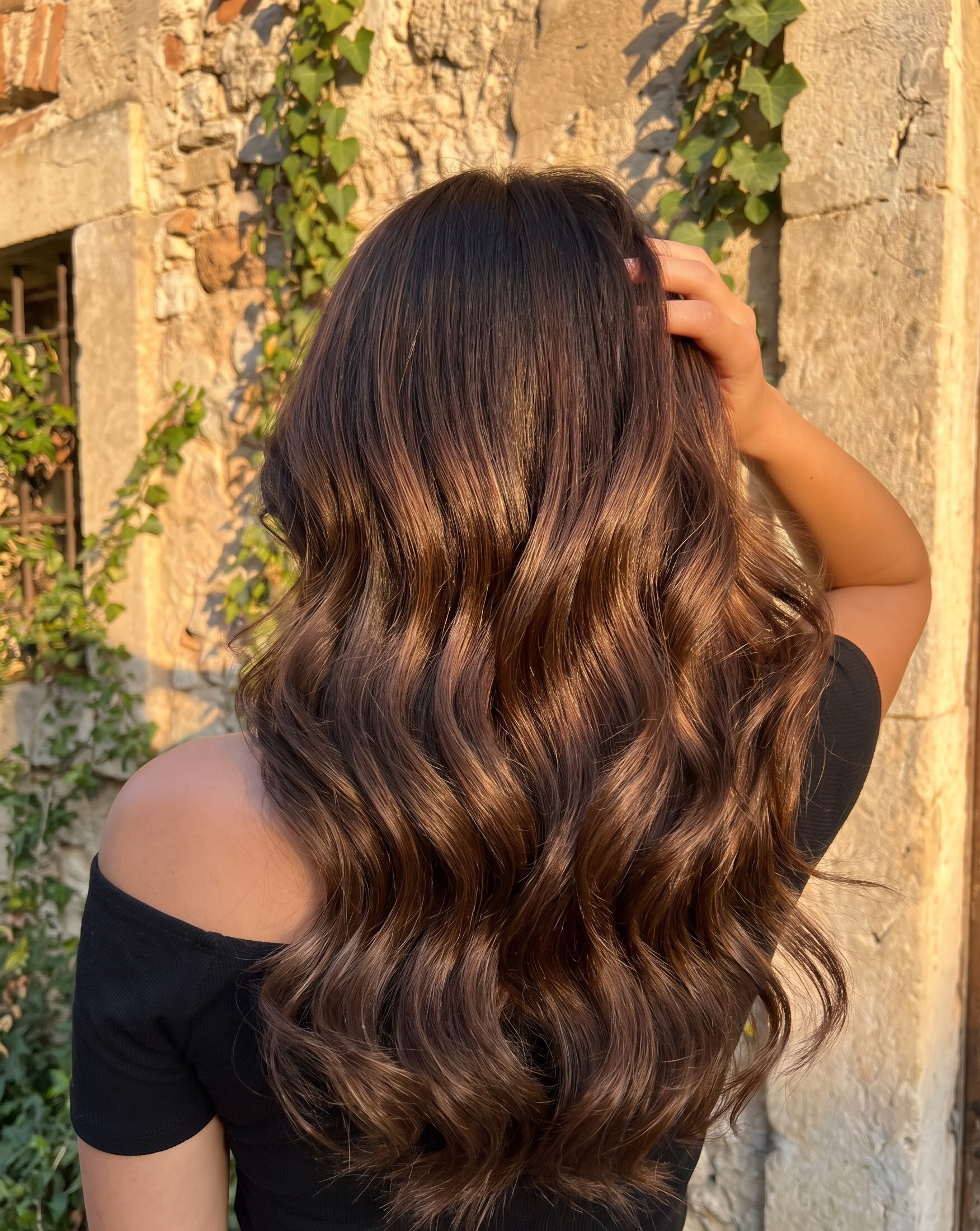 Brunette hair with caramel balayage highlights styled in loose waves against a sunlit stone wall