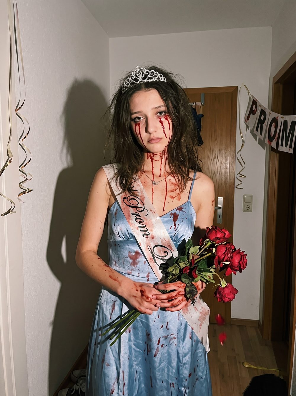 Woman in a bloody Carrie prom queen costume with a blue satin dress, a Prom Queen sash, a tiara and dead red roses