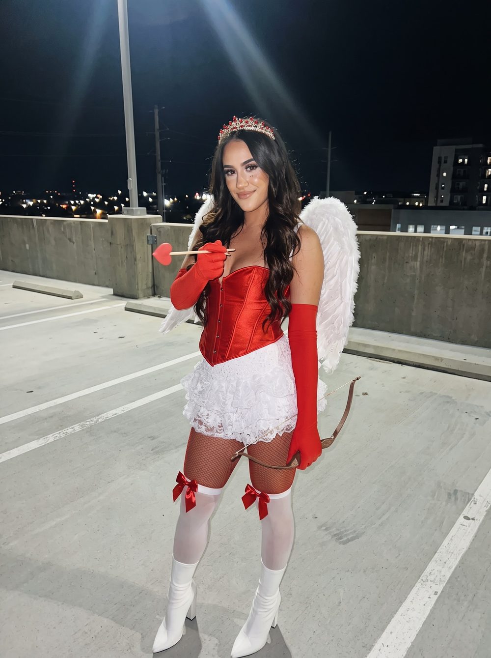 Woman in a Cupid costume with a red satin corset, white lace skirt, white feather wings, red gloves and a heart-tipped arrow