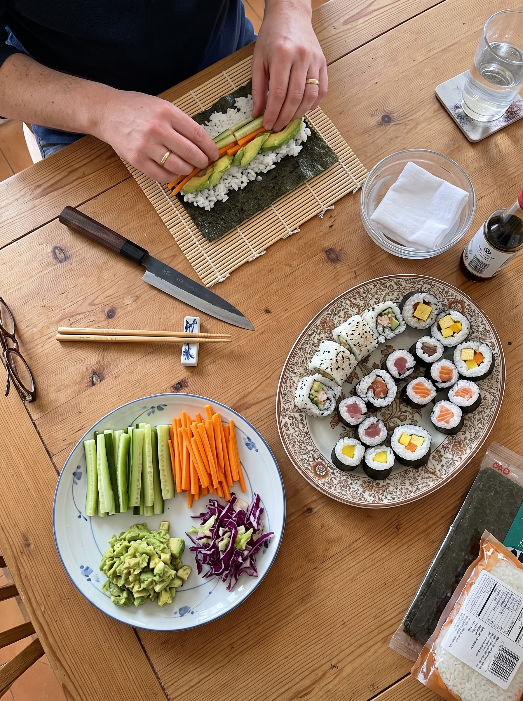 Person rolling homemade sushi with fresh vegetables on a bamboo mat next to a finished sushi platter